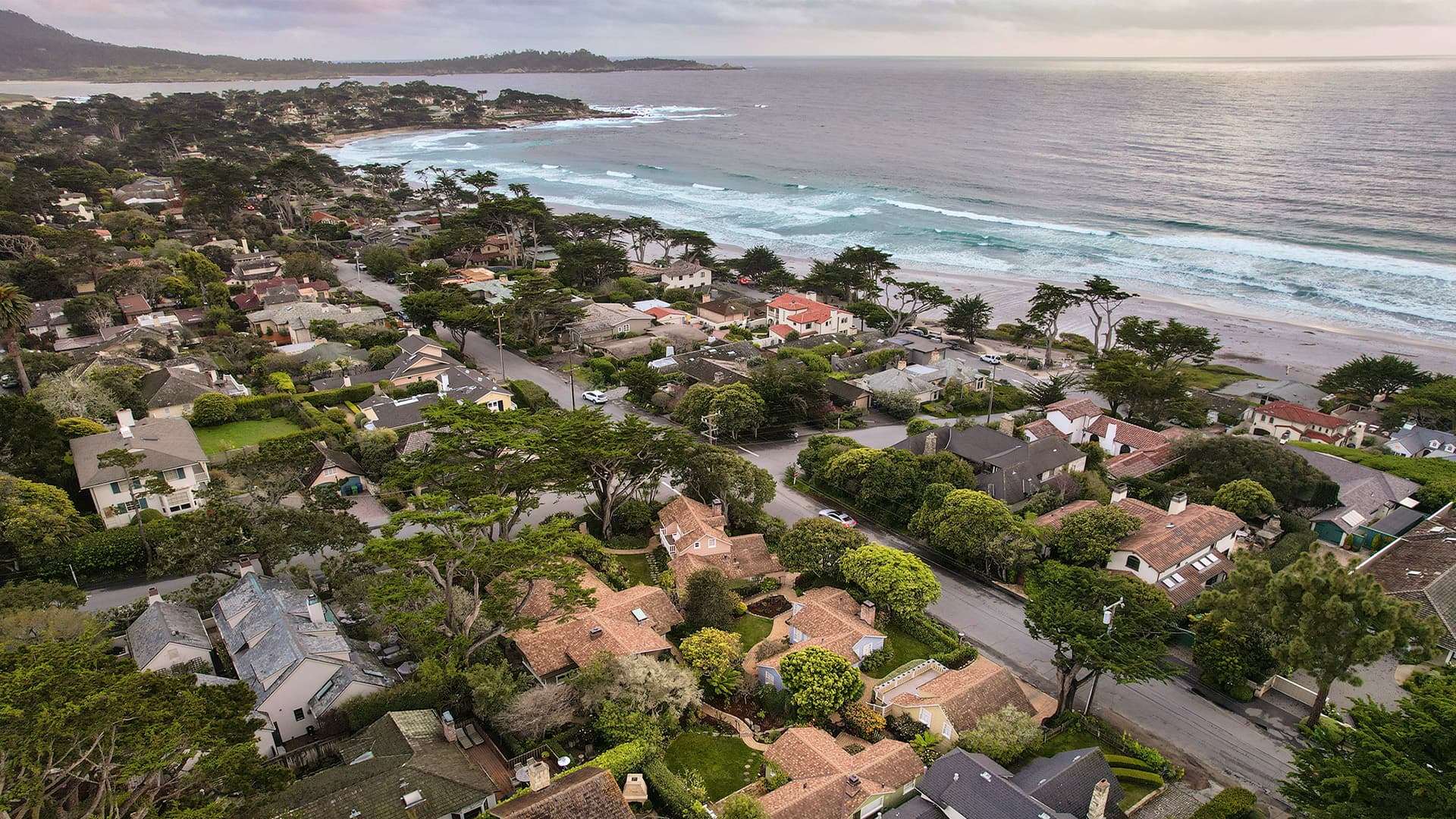 Aerial view of coastal houses and ocean waves under a cloudy sky.