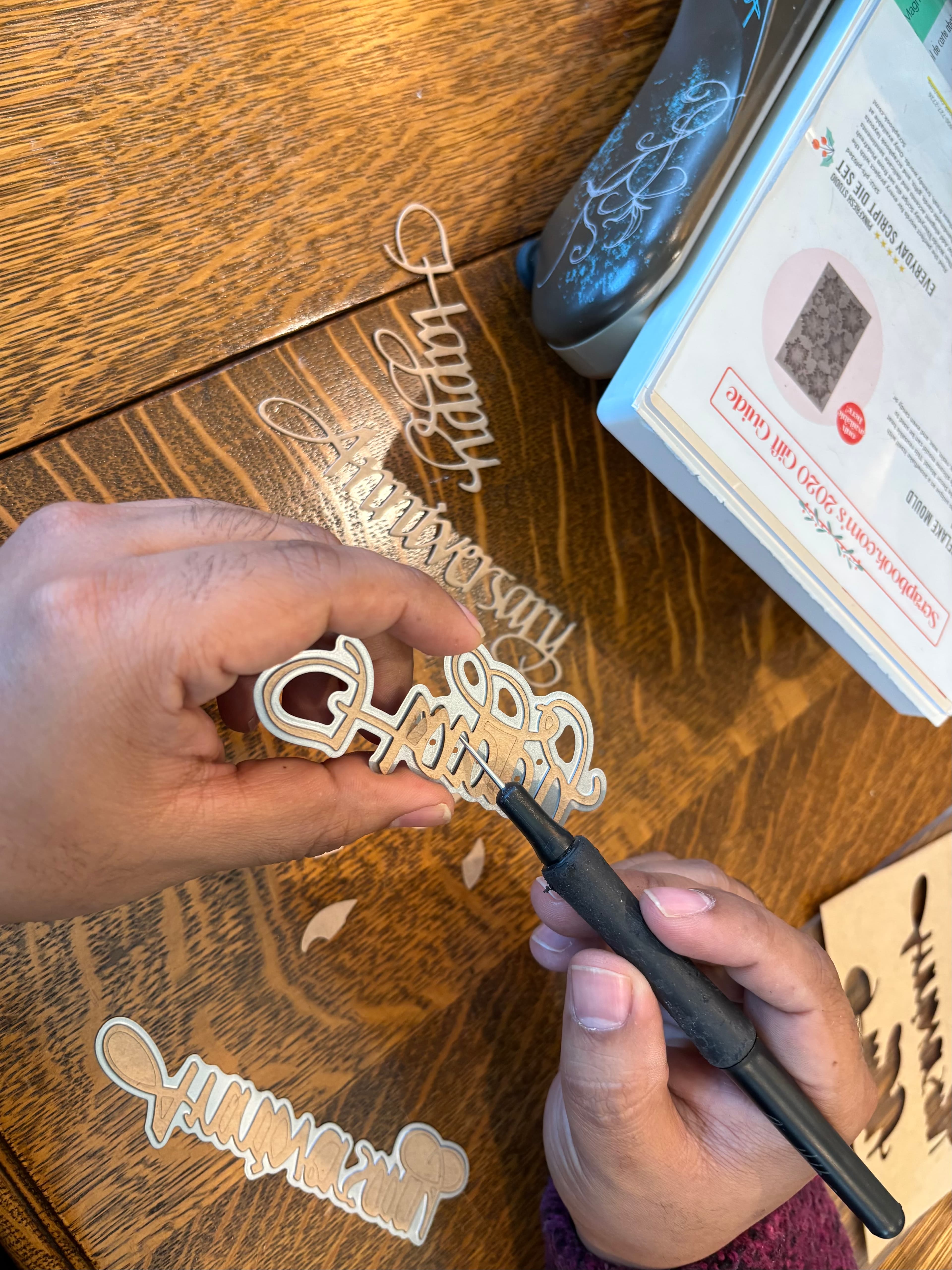A person is holding a craft tool while working on intricate paper cutouts on a wooden table.