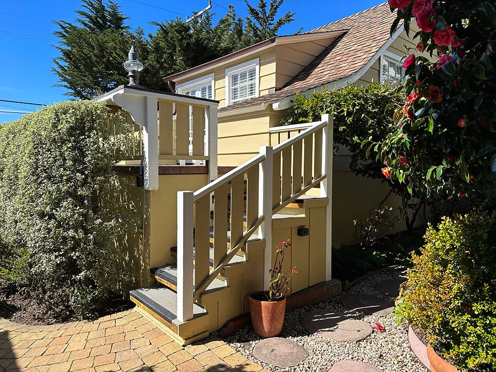 A wooden staircase leads to a house surrounded by lush greenery and colorful flowers.