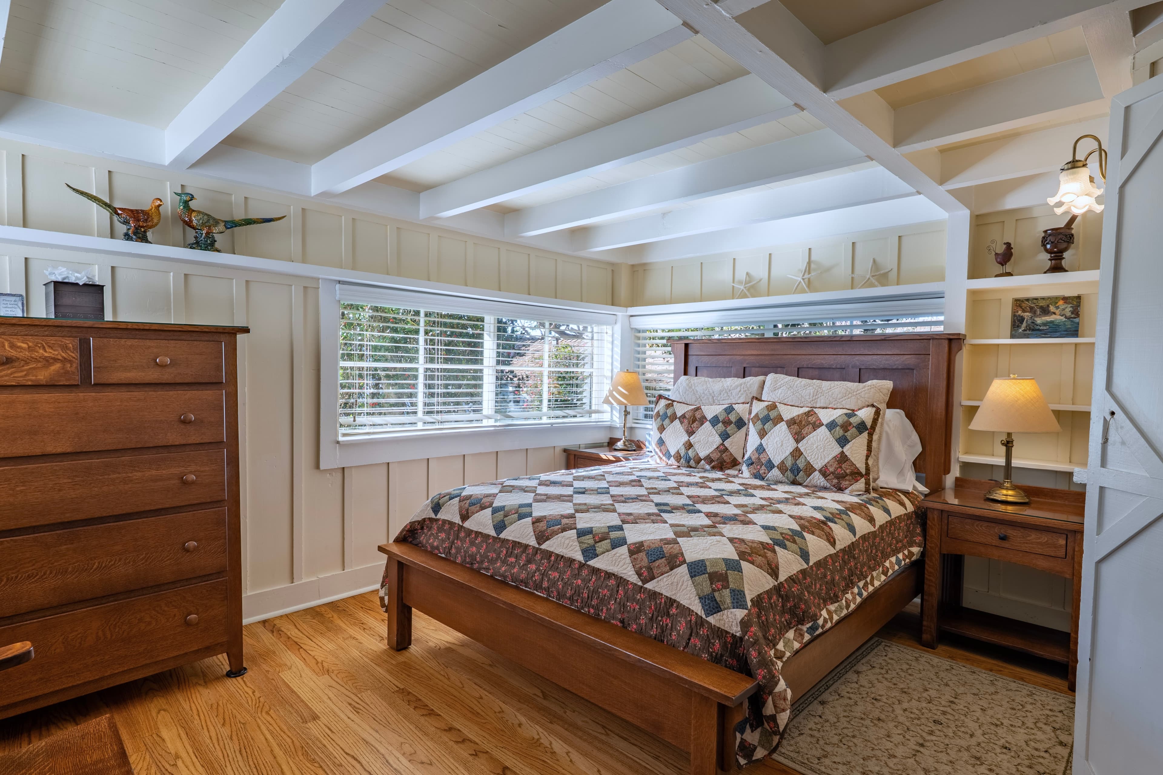 A cozy bedroom featuring a wooden bed with a quilt, matching nightstand, and dresser, illuminated by natural light from a window.