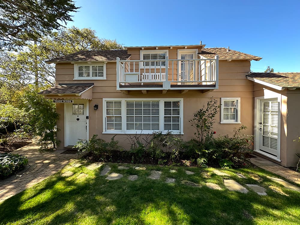 A two-story, peach-colored house with a balcony, surrounded by greenery and a stone pathway.