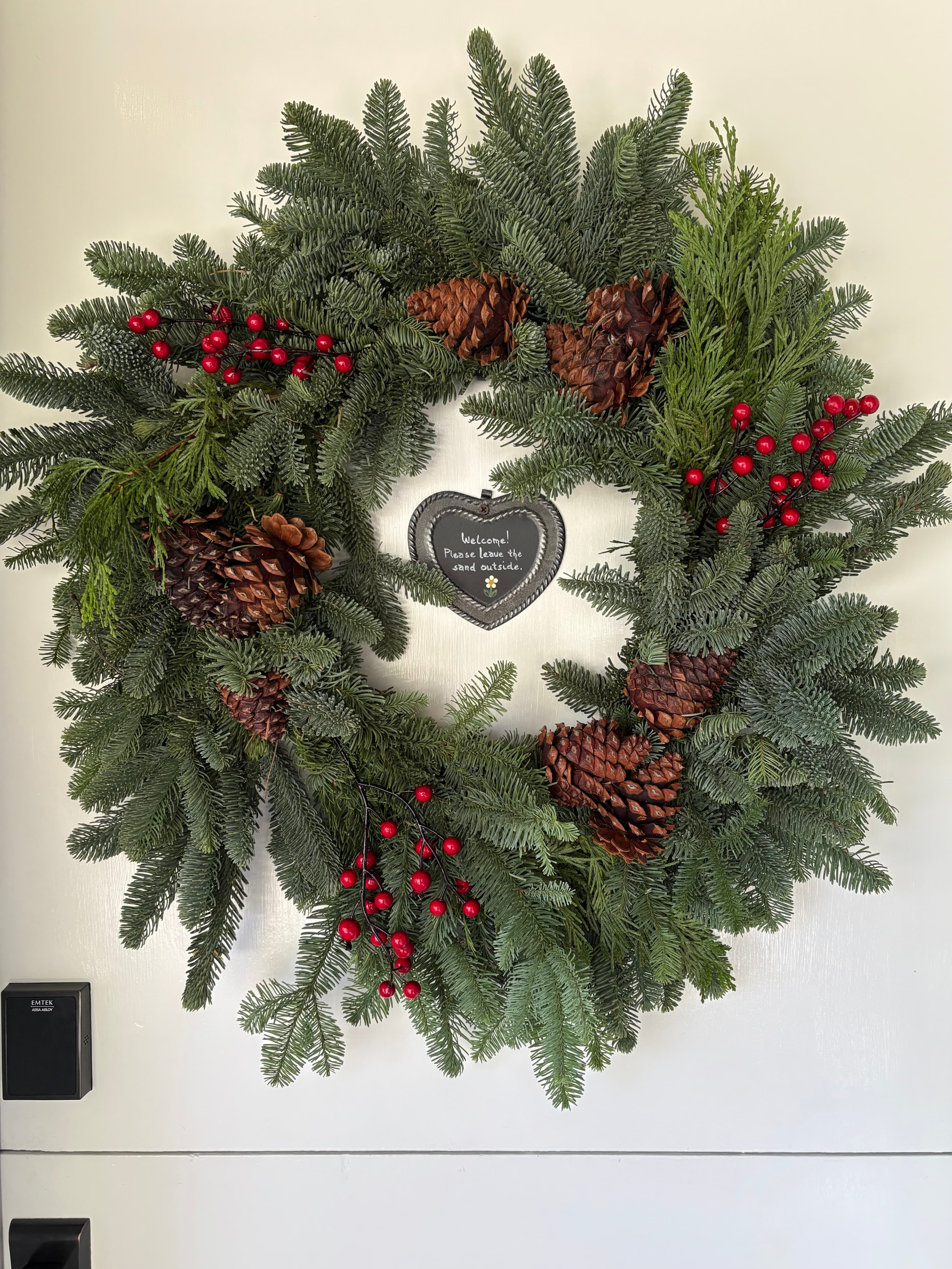 A lush green wreath adorned with pinecones and red berries, featuring a heart-shaped sign that reads "Welcome! Please leave the outside."
