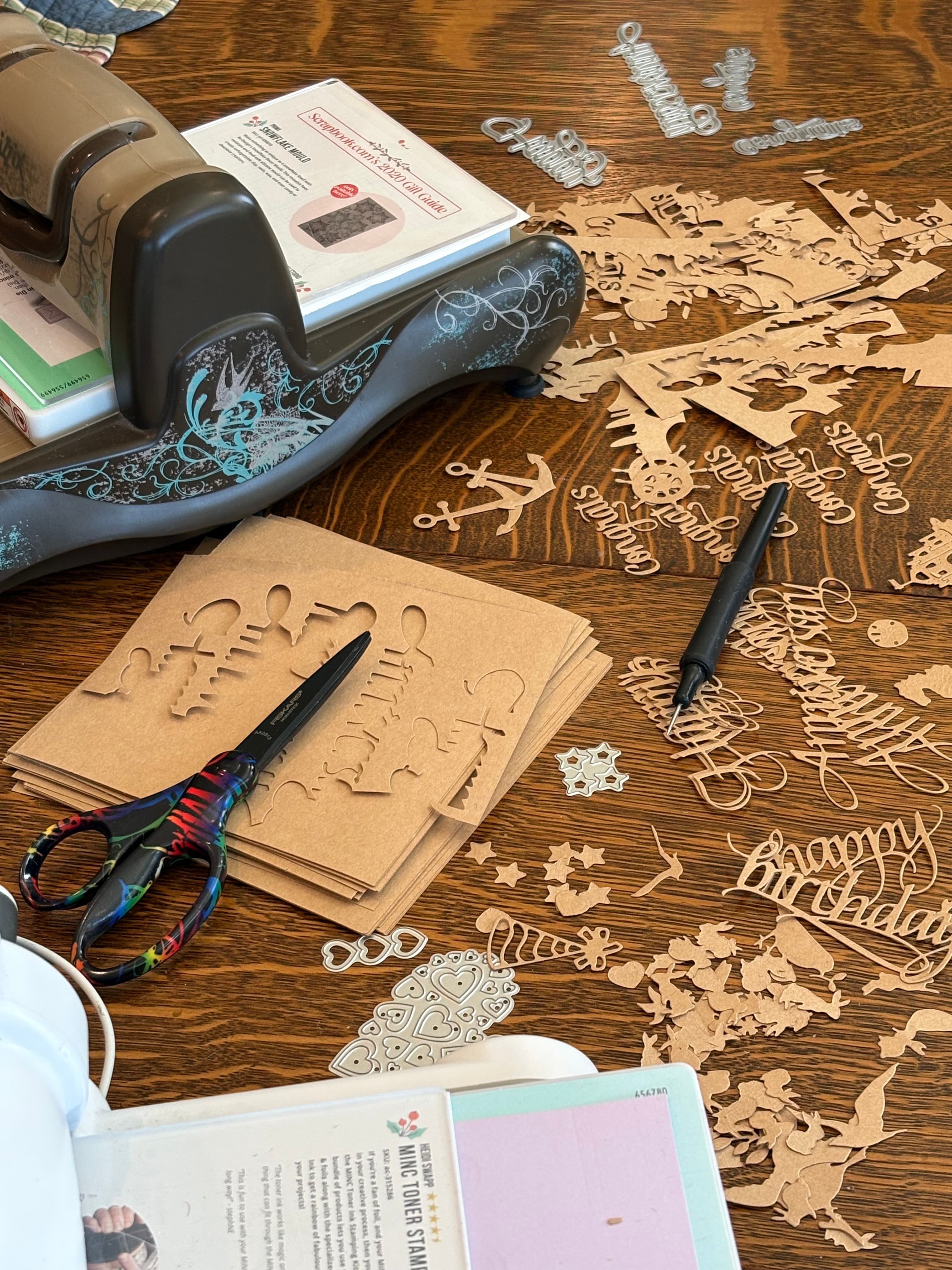 A crafting workspace features decorative cutouts, scissors, and a die-cutting machine on a wooden table.