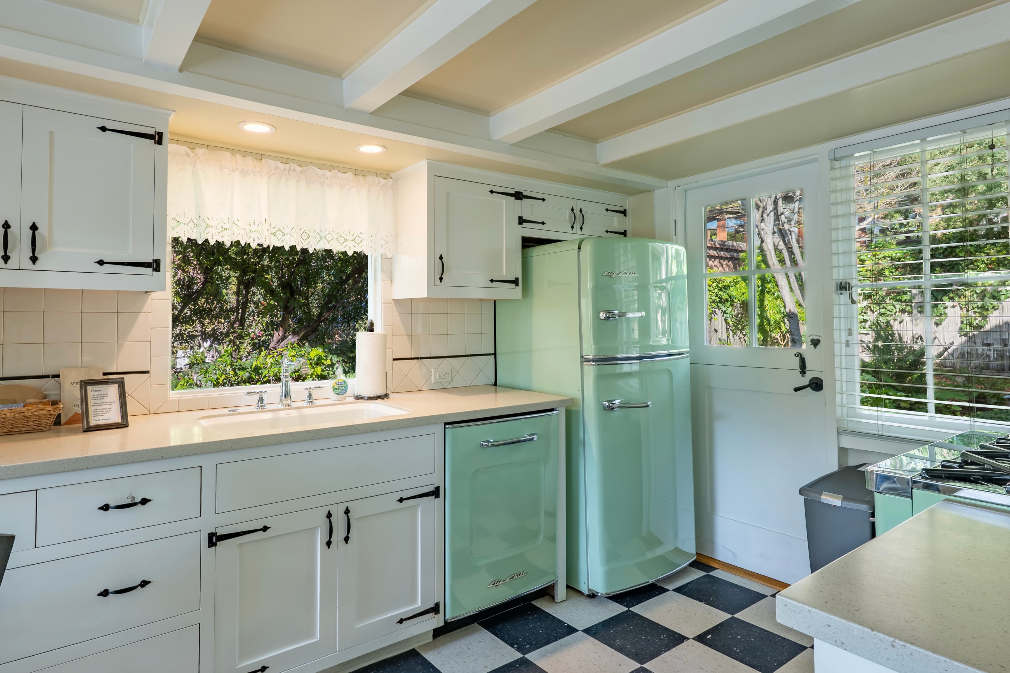 A bright kitchen featuring white cabinets, a mint green refrigerator, and a window overlooking lush greenery.
